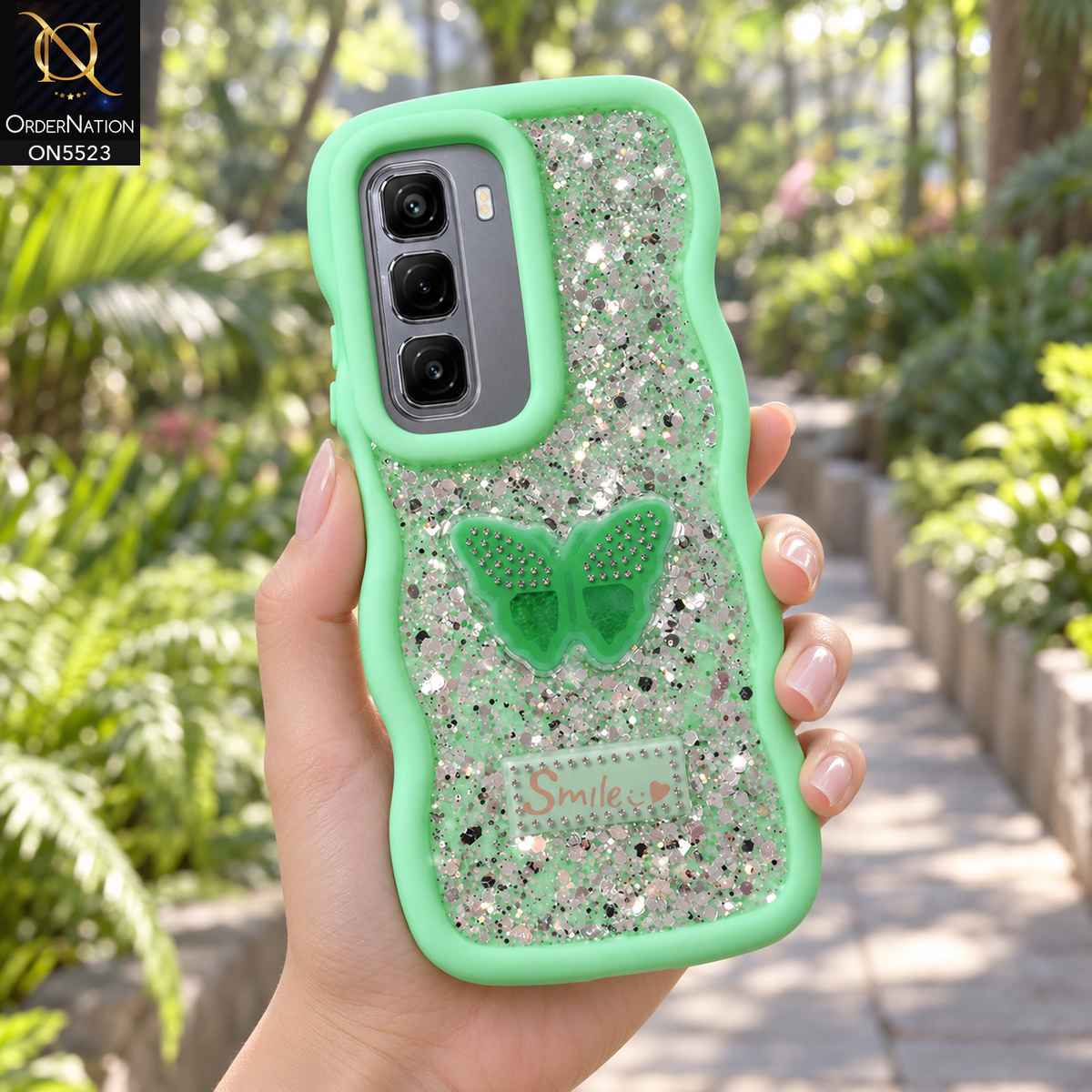 Infinix Hot 50 4G Cover - Sea Green - New Premium Smile Glitter Soft Case on the phone, held in a hand on a botanical garden path (AI lifestyle render)
